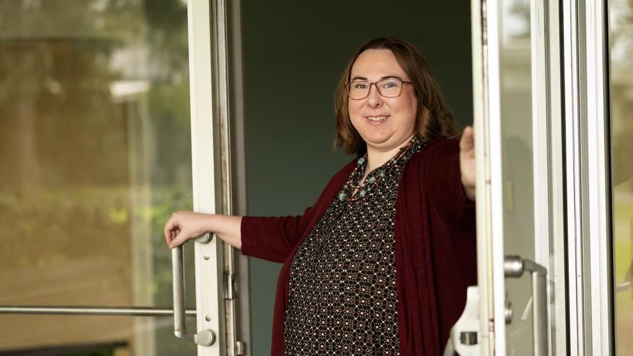 A woman with shoulder-length brown hair and glasses is standing in a doorway, holding the glass door open with both hands. She is wearing a patterned dress, a maroon cardigan, and a beaded necklace. She is smiling. The background is slightly blurred, showing an outdoor scene with greenery and reflections in the glass.