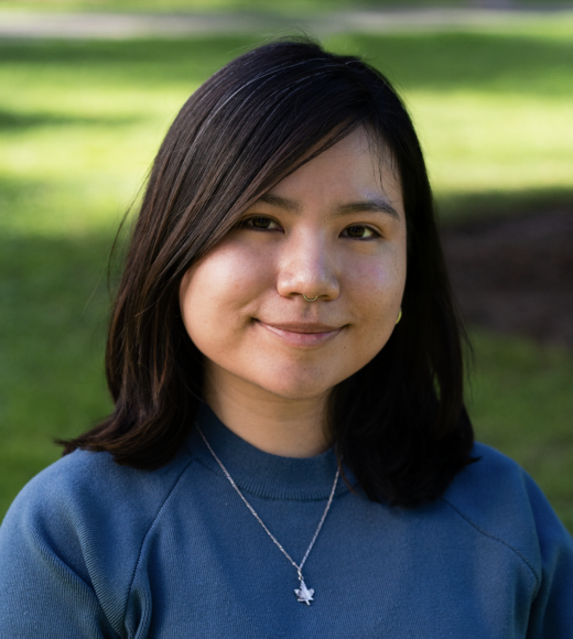 Smiling woman with shoulder-length dark hair, wearing a blue sweater, in a park with green grass.