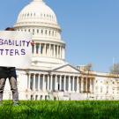 A person stands on the grass in front of the United States Capitol building holding a large white sign that reads “Disability Matters.”